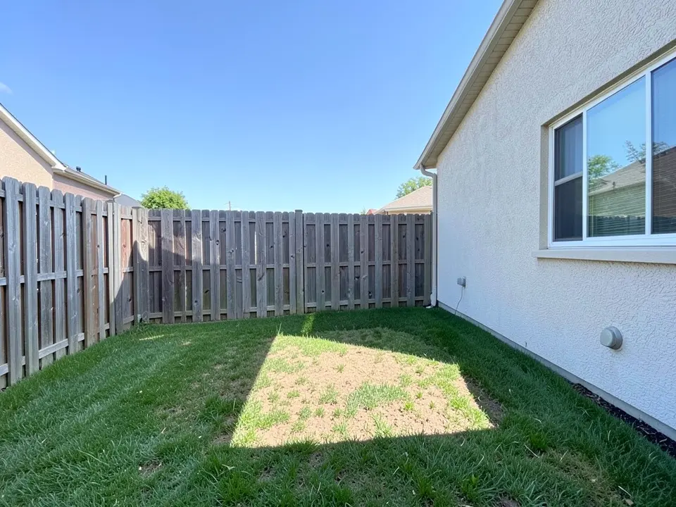 AI-generated example: a corner of a backyard with a grey weathered wooden fence, the cream stucco side of a house with a large window and a grey box on the wall, a mown lawn with a bare, thin sunlit patch in the middle, under a blue sky