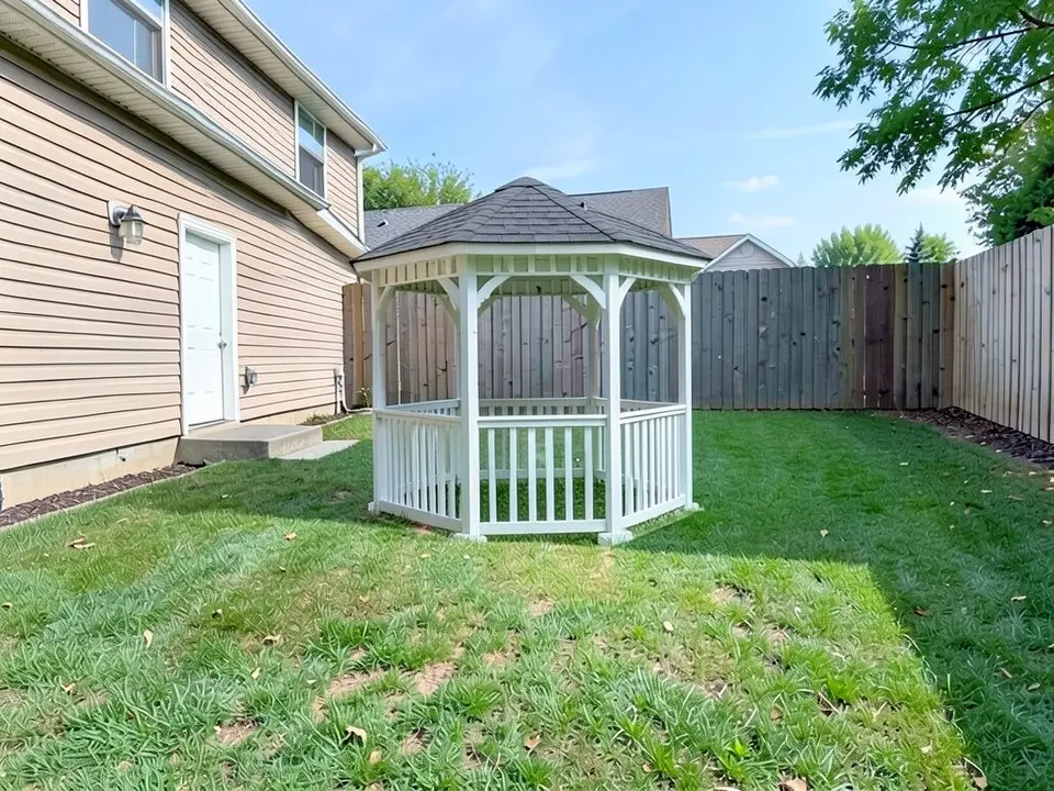 AI-edited version of the same image: a white wooden gazebo with a dark shingle roof, railings and open sides stands on the lawn in front of the back fence