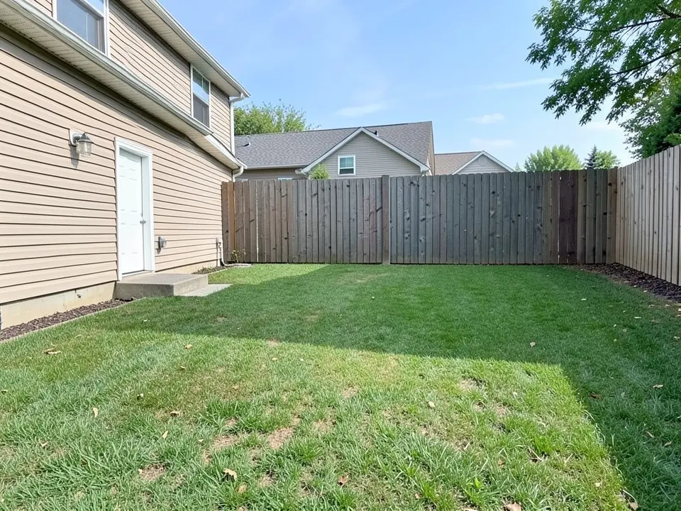 AI-generated example: a plain green backyard lawn beside a beige two-storey house with a white side door, a tall wooden fence along the back and nothing on the grass