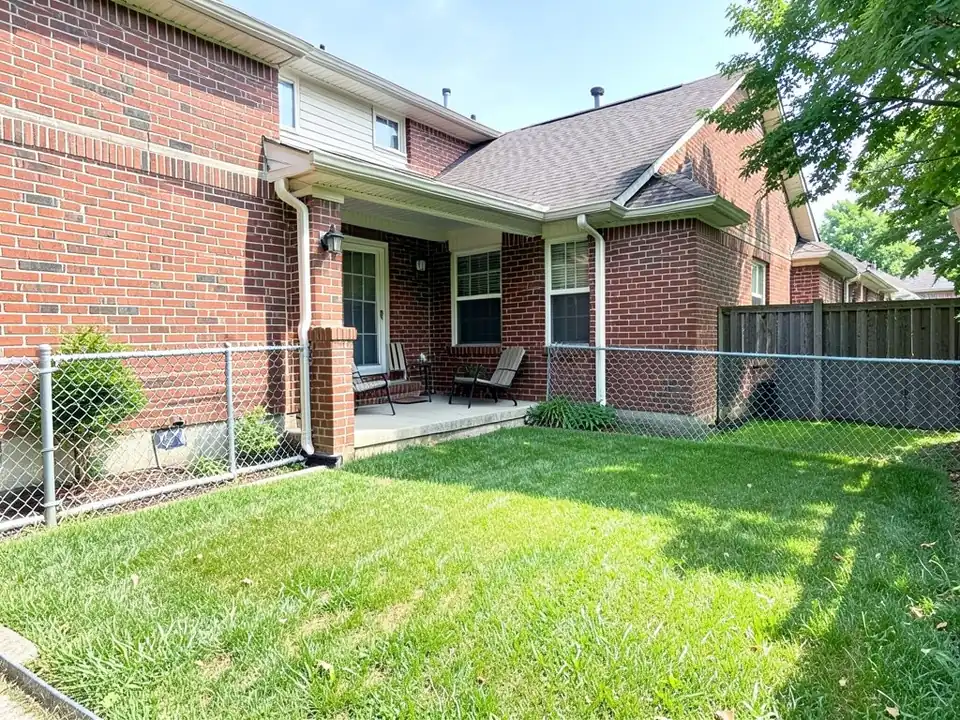 AI-generated example: a small lawn behind a red brick house with a covered patio, two patio chairs and a small table, a chain link fence and a dark wooden fence beyond it