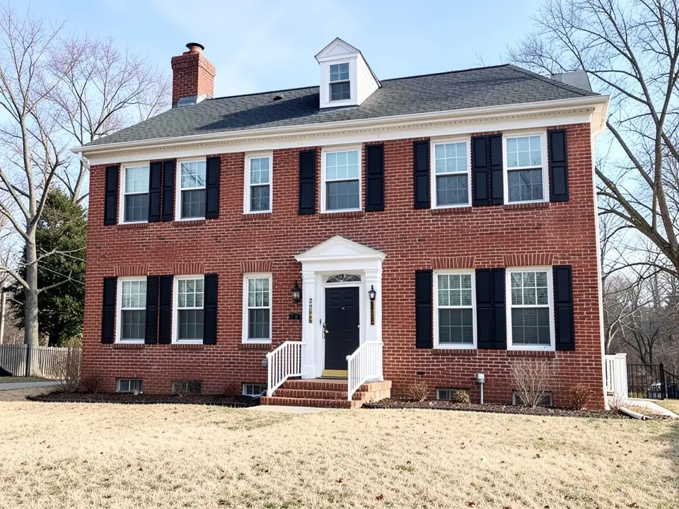 AI-generated example: a red brick two-story colonial house with white trim, black shutters, a white columned entry with white stair railings, a dormer and a chimney, thin strips of dark mulch along the foundation with a few small bare shrubs, a dry tan lawn and bare winter trees