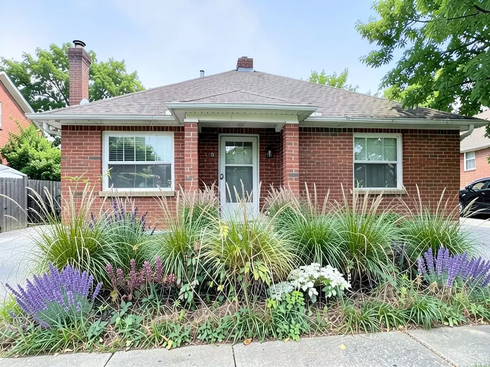 AI-edited version of the same image: the hedge is gone and a bed of tall ornamental grass clumps runs across the front of the brick house, with purple spiky flowers at both ends, a few pink spikes and a clump of white flowers in the middle, and the lower brick wall, the door step and pale paving at both ends now visible