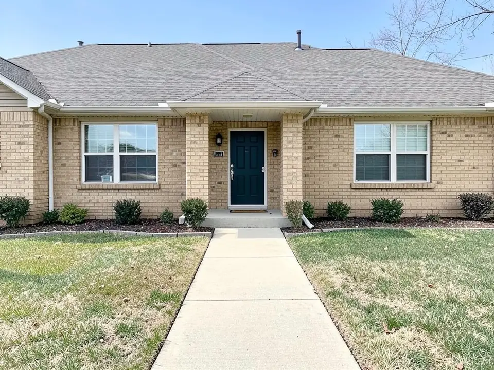 AI-generated example: a one-story tan brick house with a dark green front door under a small gabled porch, two white windows, a narrow mulch bed with small shrubs along the foundation, and a straight concrete walkway across a patchy lawn