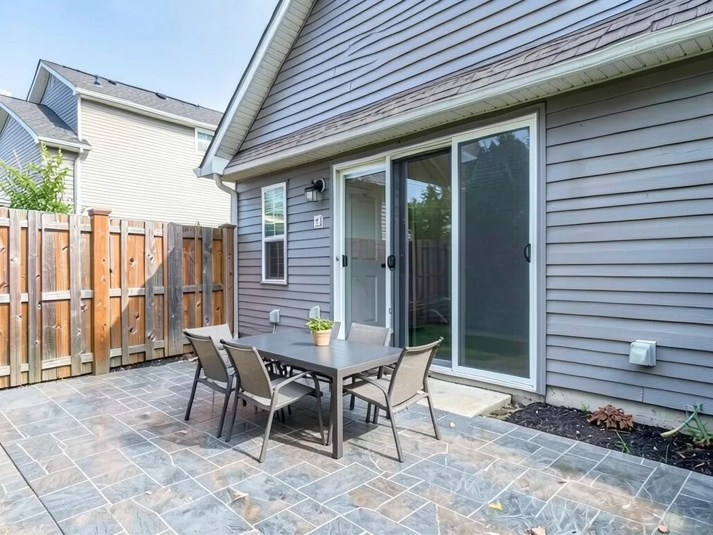 AI-edited version of the same image: a blue-grey stamped concrete patio in a stone pattern where the lawn was, with a rectangular dining table, six chairs and a potted plant on the table