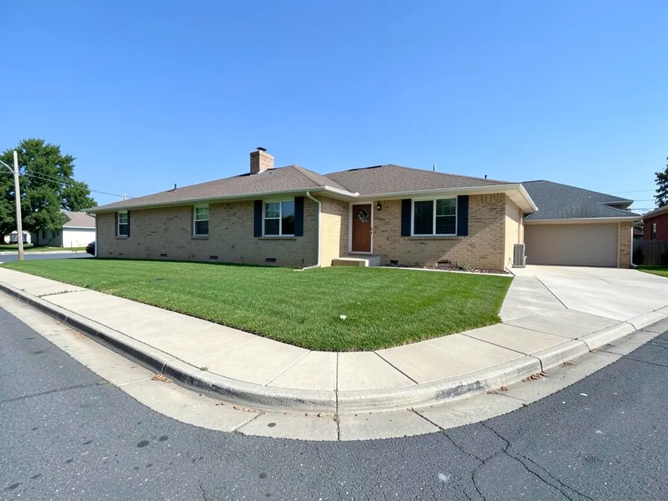 AI-generated example: a single-story tan brick ranch house with navy shutters on a corner lot, a plain green lawn across the front and around the left side, a concrete sidewalk that turns the street corner in the foreground and a concrete driveway to an attached garage on the right