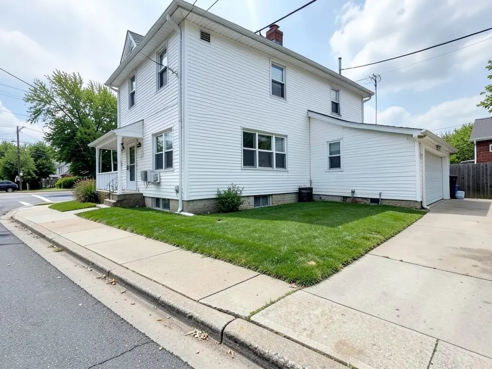 AI-generated example: the long side wall of a white two-story corner house with a small front porch on the left, a strip of plain lawn between the wall and a concrete public sidewalk, the street intersection in the distance on the left and a concrete driveway to a garage on the right