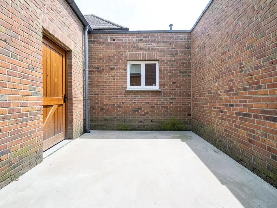 AI-generated example: a small courtyard with a plain pale concrete floor and tall red brick walls on three sides, a wooden door on the left, a small white-framed window in the back wall, a grey drainpipe in the corner, and some moss on the lower brick, in daylight