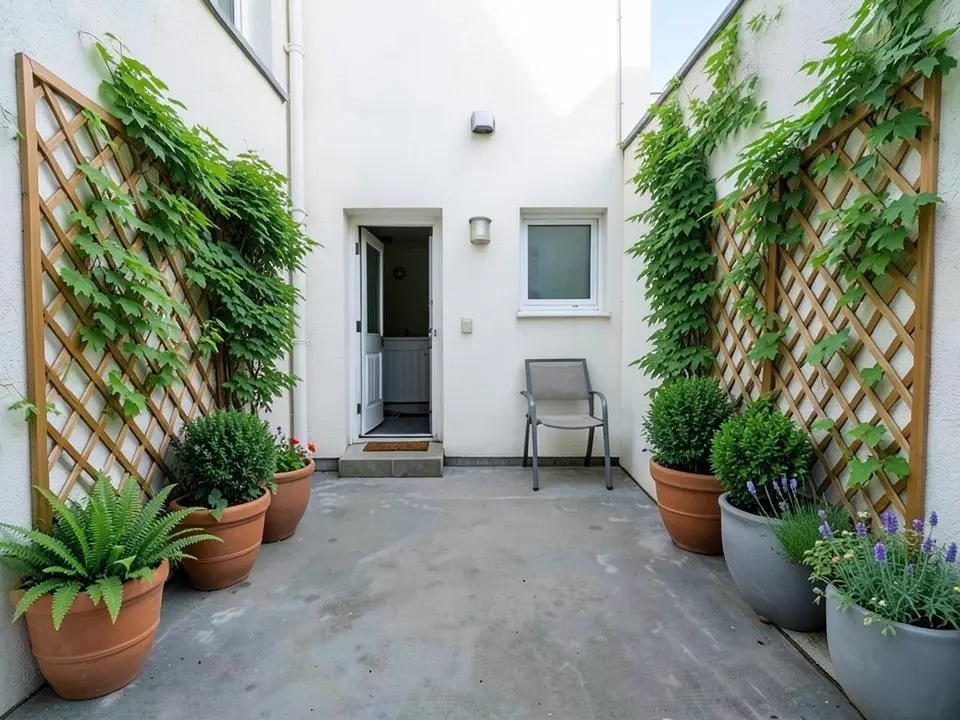 AI-edited version of the same image: a wooden lattice trellis with green climbing leaves on each side wall, terracotta pots with a fern, a round green shrub and flowers on the left, and terracotta and grey pots with round shrubs and lavender on the right, with the door, the window, the chair and the paving as before