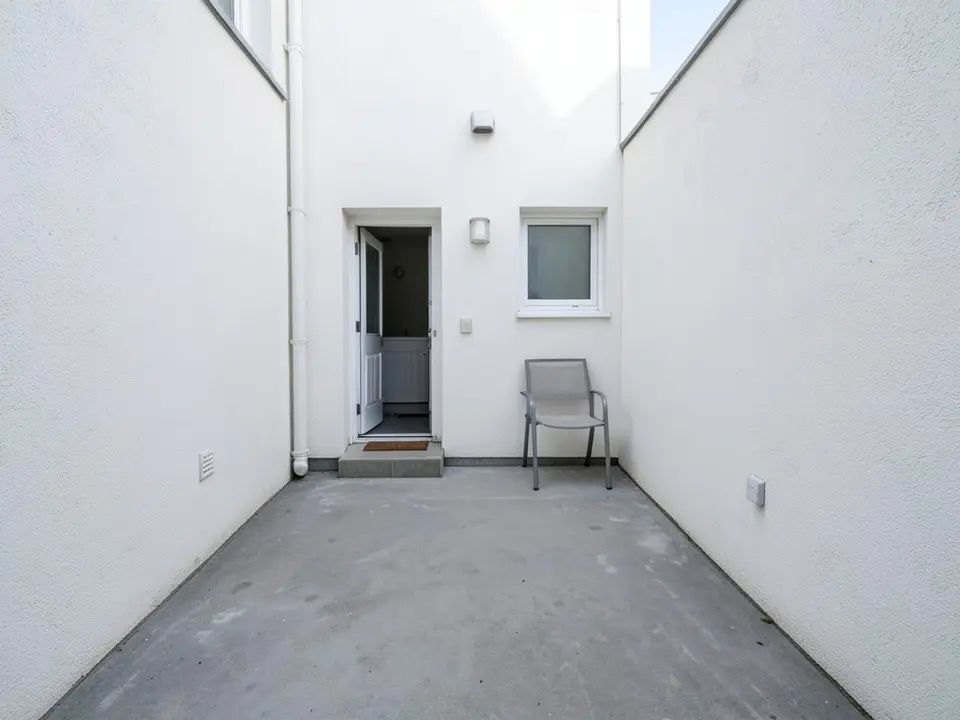 AI-generated example: a narrow courtyard with white rendered walls on both sides and at the back, grey concrete paving, a white back door standing open with a concrete step and a doormat, a small window, two wall lights, a white drainpipe, one grey metal chair against the back wall, a vent on the left wall and a socket on the right wall, in daylight