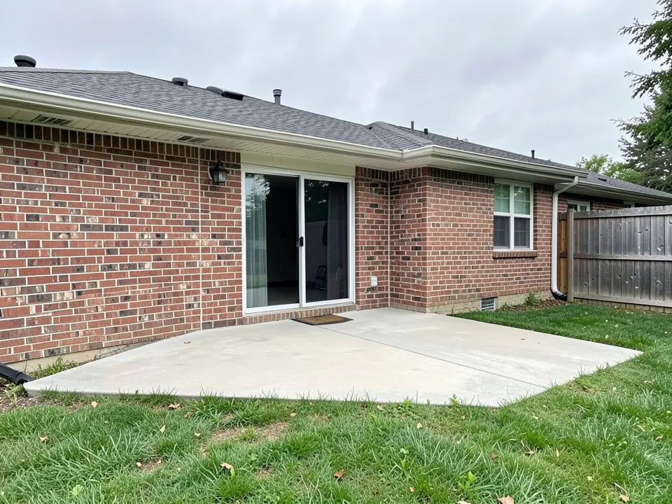 AI-generated example: the back of a one-story red-brick house with a sliding glass door and a doormat, a plain concrete patio slab, a green lawn and a wooden fence on the right