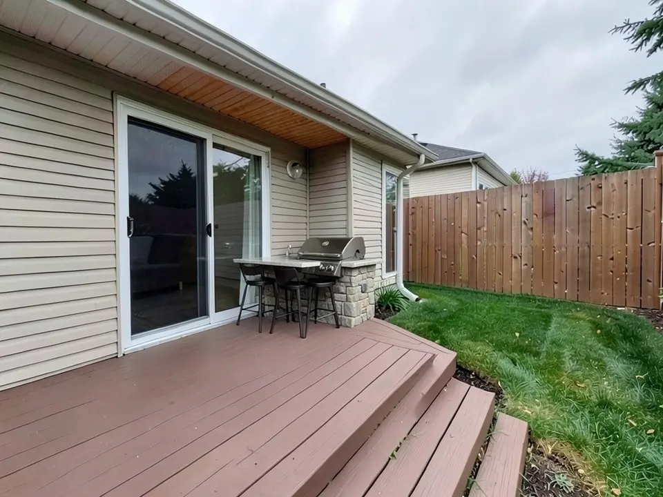 AI-edited version of the same image: a small stone-faced counter with a built-in grill and three bar stools stands on the deck by the door, and the underside of the house eave above it is now wood-colored