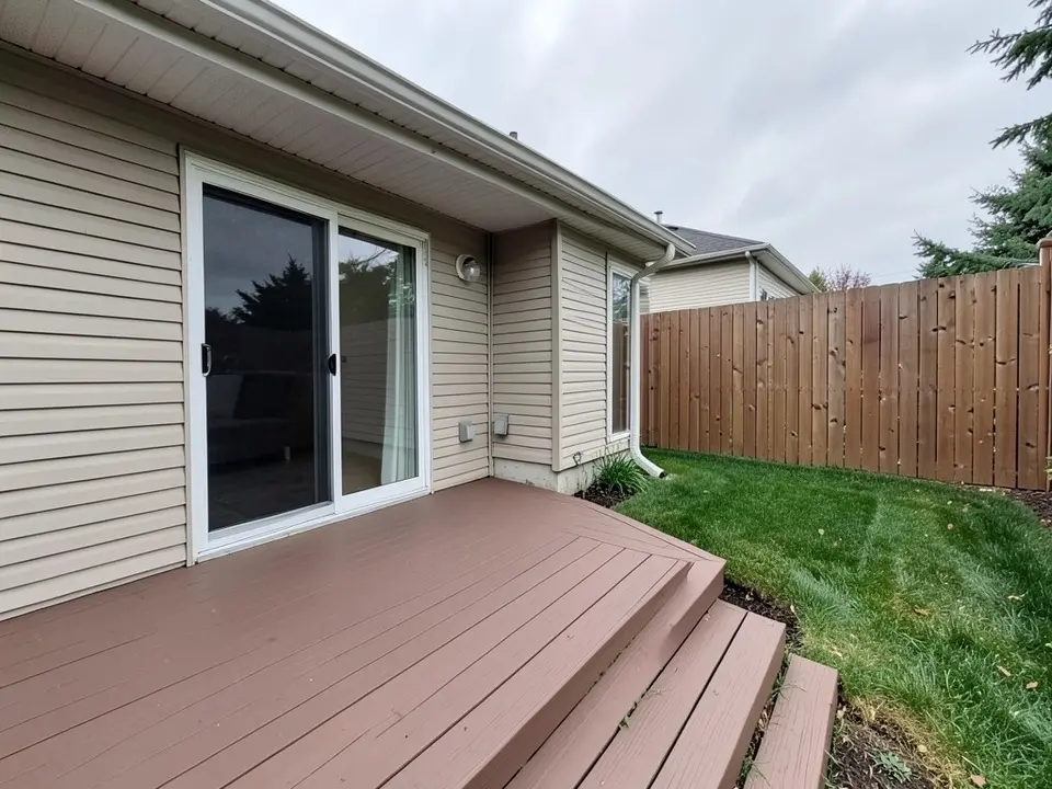 AI-generated example: the corner of a beige house with a sliding glass door, a plain brown composite deck with steps in front, a lawn and a tall wooden fence