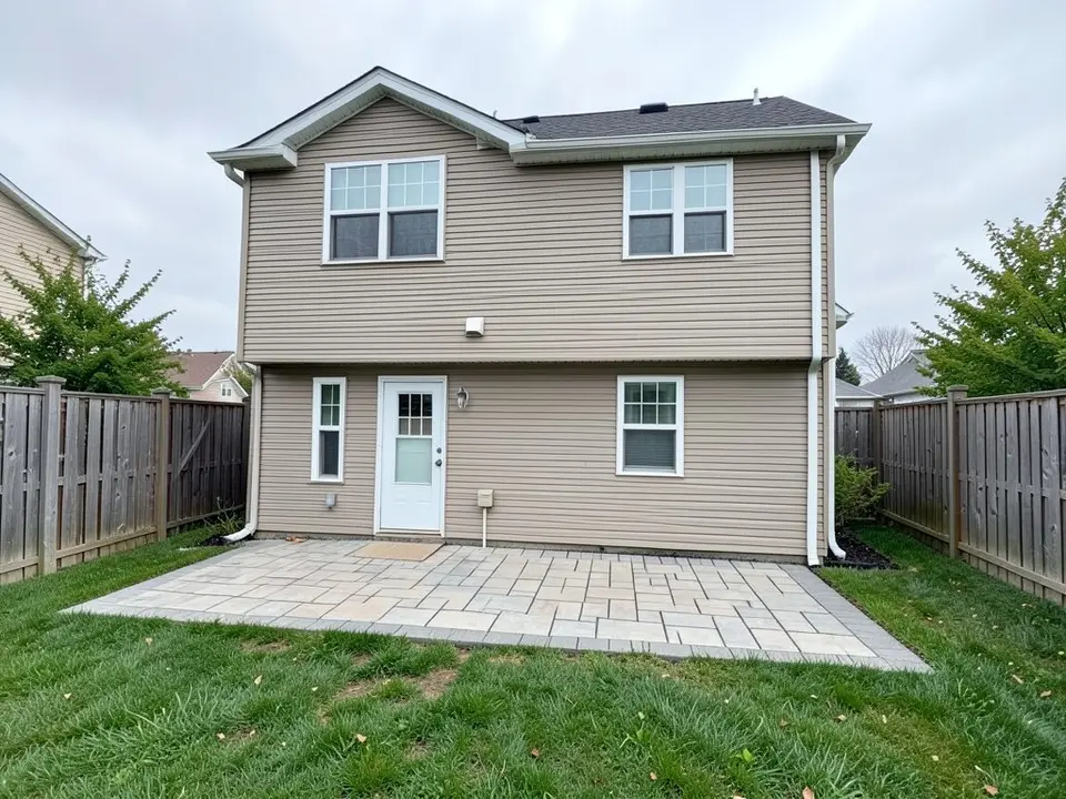 AI-generated example: the back of a two-story house with beige siding, a door and windows, a patio of light stone pavers on the ground, a green lawn and wooden fences on both sides
