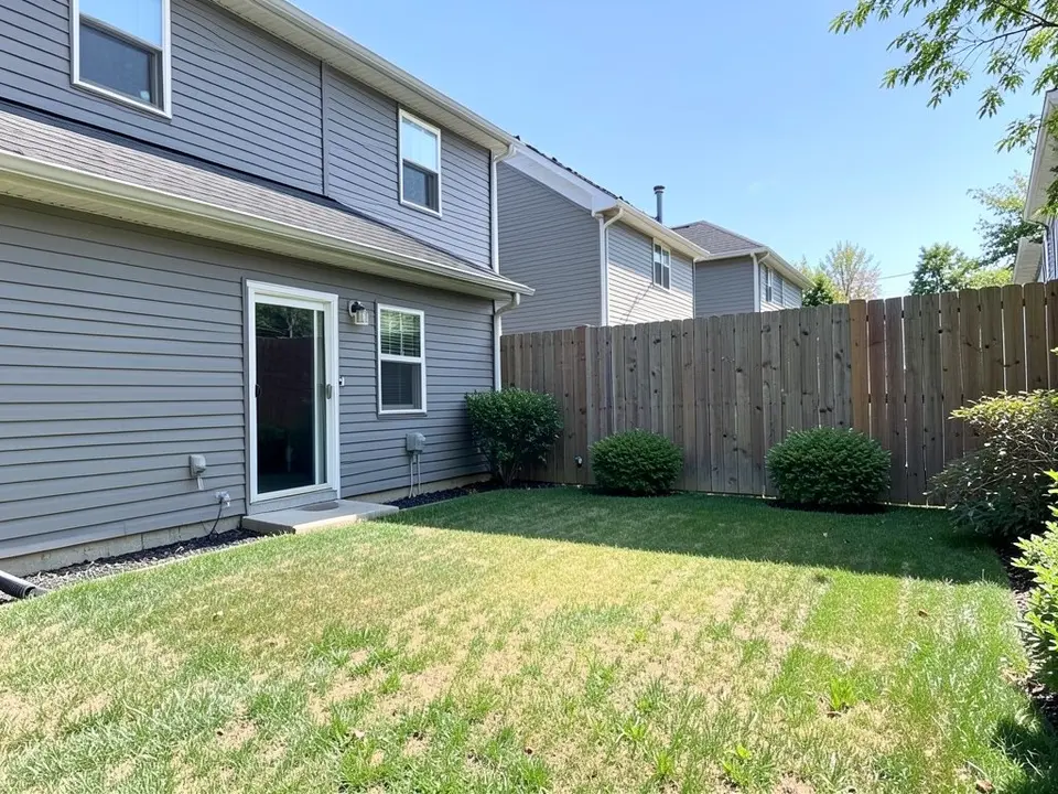 AI-generated example: the back of a grey two-story house with a sliding patio door and a lawn with dry patches running up to the house wall, three round shrubs along a tall wooden fence and a strip of black mulch beside the wall, in bright sun