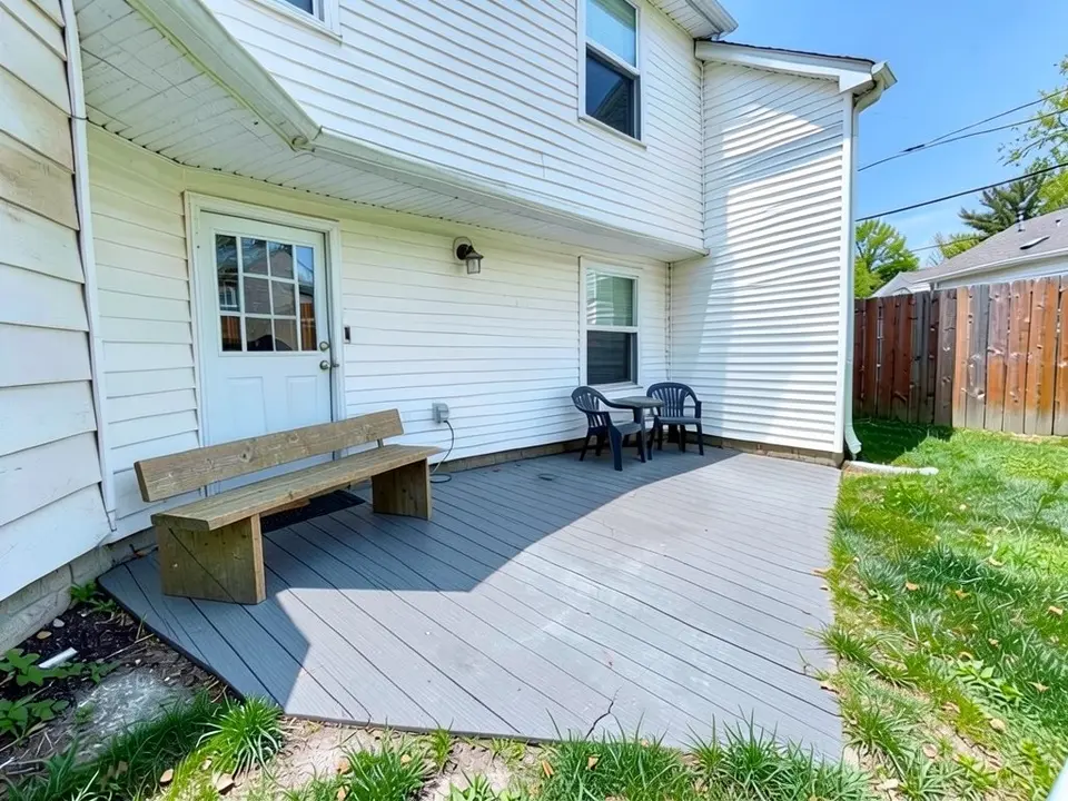 AI-edited version of the same image: the slab area covered with grey boards and a long plain wooden bench with a back standing against the house wall beside the door, with the chairs and table still in place and the doormat mostly hidden behind the bench