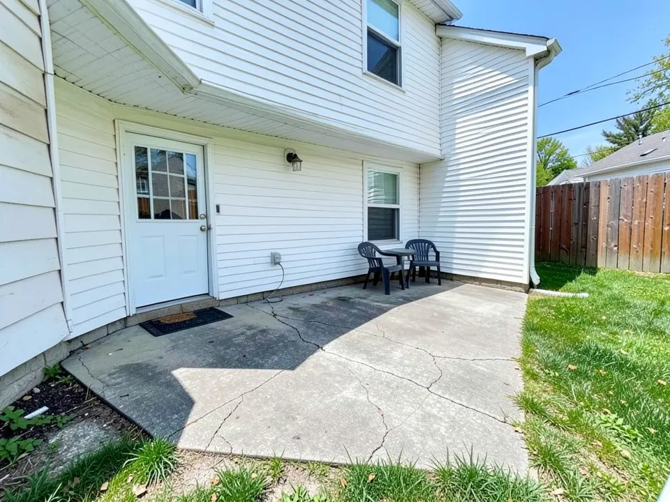 AI-generated example: a small cracked grey concrete slab at the back door of a white two-story house, two dark chairs and a small table in the corner by the right-hand wall, a doormat at the door, a wooden fence and a strip of lawn on the right under a blue sky