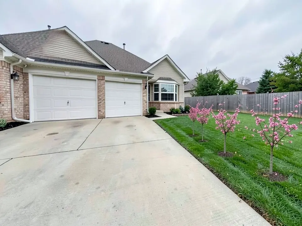 AI-edited version of the same image: four small trees with pink blossoms in an uneven row along the lawn next to the driveway, each standing on a small ring of dark mulch, with the house, driveway and fence otherwise as before