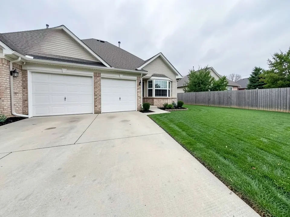 AI-generated example: a brick and beige house with a two-car garage with white doors, a bay window, a wide concrete driveway at the front and a wide green lawn to its right running up to a wooden fence, under an overcast sky