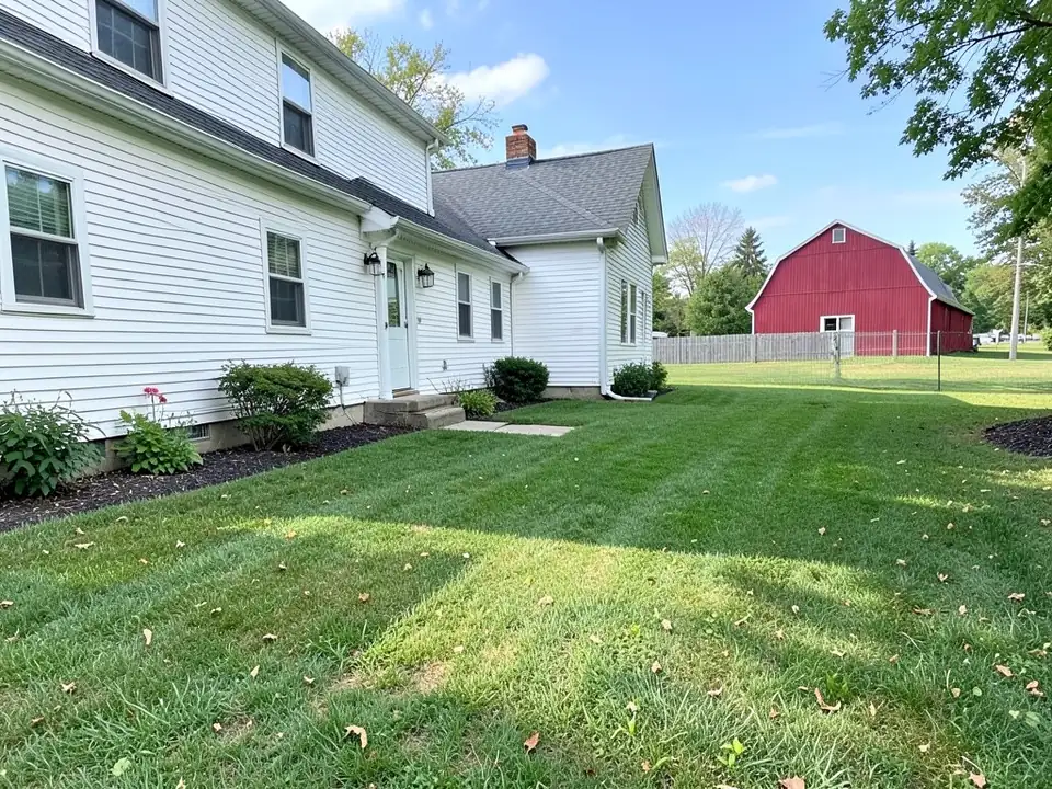 AI-generated example: the side yard of a white two-story farmhouse with a side door and a concrete step, shrubs in dark mulch along the house, a striped mowed lawn with scattered leaves, and a red barn behind a wire fence in the distance
