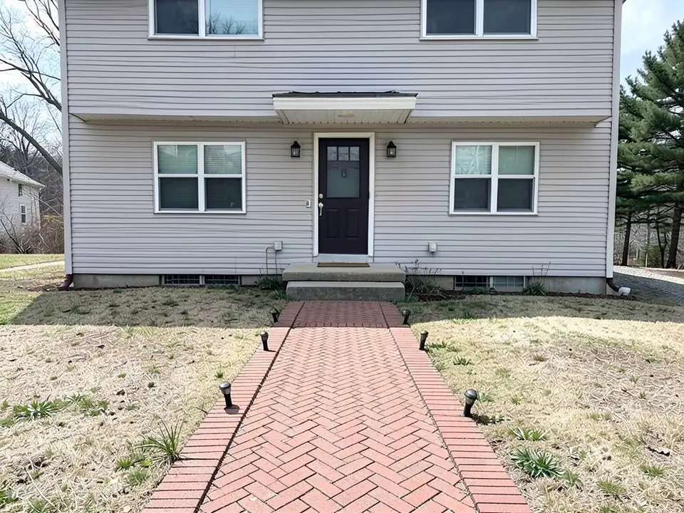 AI-edited version of the same image: a wider red brick walkway in a herringbone pattern with a band of crosswise bricks along each edge and six small black stake lights in the grass beside it