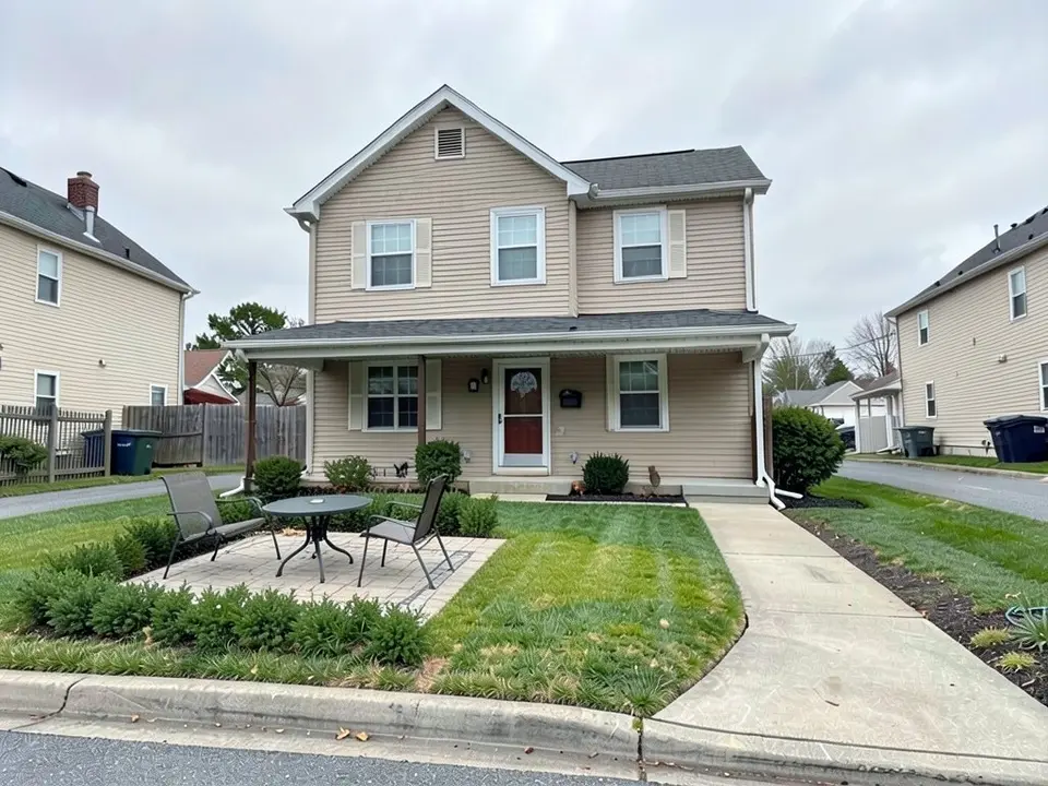 AI-edited version of the same image: a small square paver patio with a round table and two metal chairs on the left side of the lawn, with a row of low green shrubs in front of it and to its left, and a thicker, greener lawn to the right of it