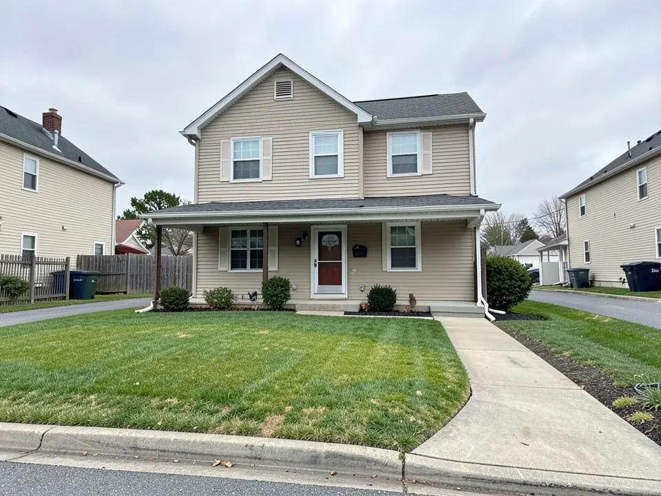 AI-generated example: a beige two-story house with a covered porch and a red front door, a curved concrete walkway, a mown lawn with a few thin yellowish patches running to the street curb, neighbors' houses and trash bins on both sides, under an overcast sky