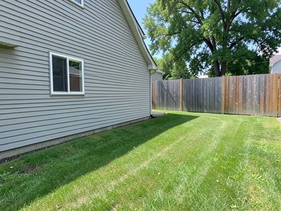 AI-generated example: a flat strip of green lawn beside the grey vinyl siding wall of a two-story house with one full window, a tall weathered wooden fence at the far side and a large tree behind the fence