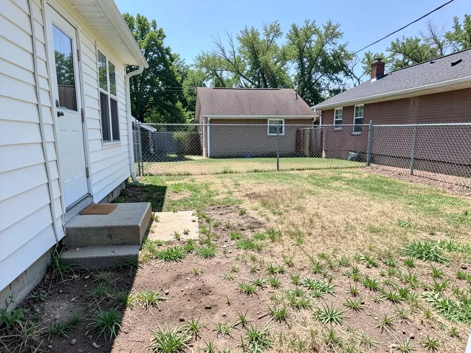 AI-generated example: a small backyard with a thin, patchy lawn, bare dirt and weeds, a short concrete step and a flat concrete pad at the back door of a white house on the left, a chain-link fence across the back, a garage and a brick house beyond it and tall trees