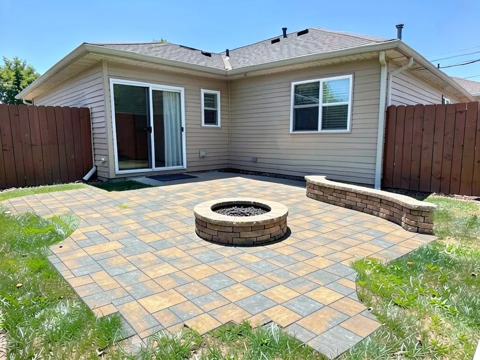 AI-edited version of the same image: a large part of the lawn is now a patio of tan and grey square concrete pavers with a round stone fire pit in the middle and a low curved stone seat wall at the back right; the small concrete slab and doormat are still by the door, and strips of lawn remain at the sides