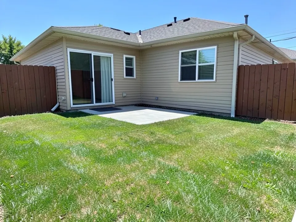 AI-generated example: the back of a beige one-story house with a sliding glass door, a small window and a larger window, white downspouts, a small plain concrete slab with a doormat outside the sliding door, brown wooden privacy fences on both sides and a green lawn in front