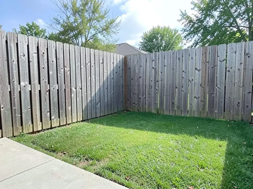AI-generated example: the back corner of a suburban backyard where two tall weathered wooden privacy fences meet, with a plain green lawn and the edge of a concrete patio at the bottom left