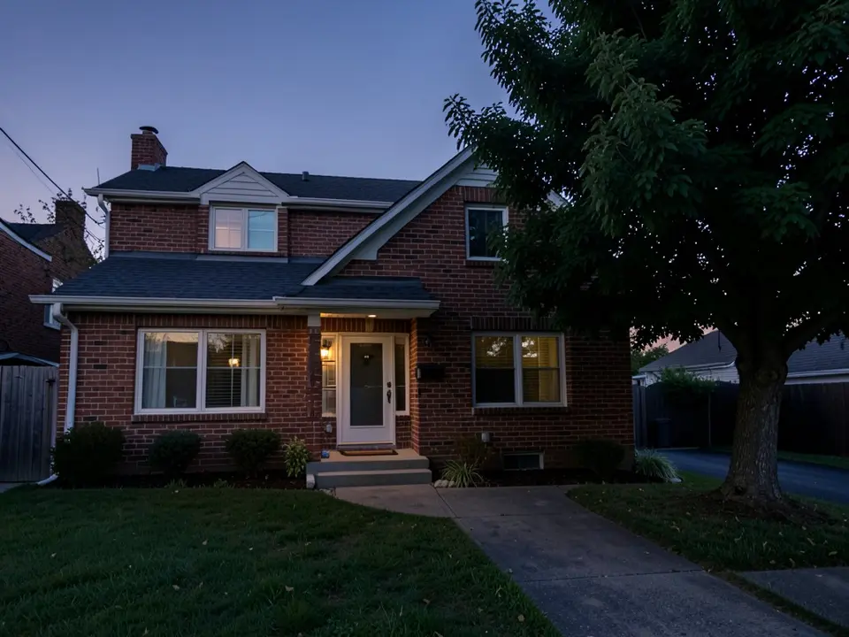 AI-generated example at dusk: a two-storey red brick house with a white front door, lit windows and a large tree on the right, with a concrete walkway and lawn and no outdoor lights on the walls