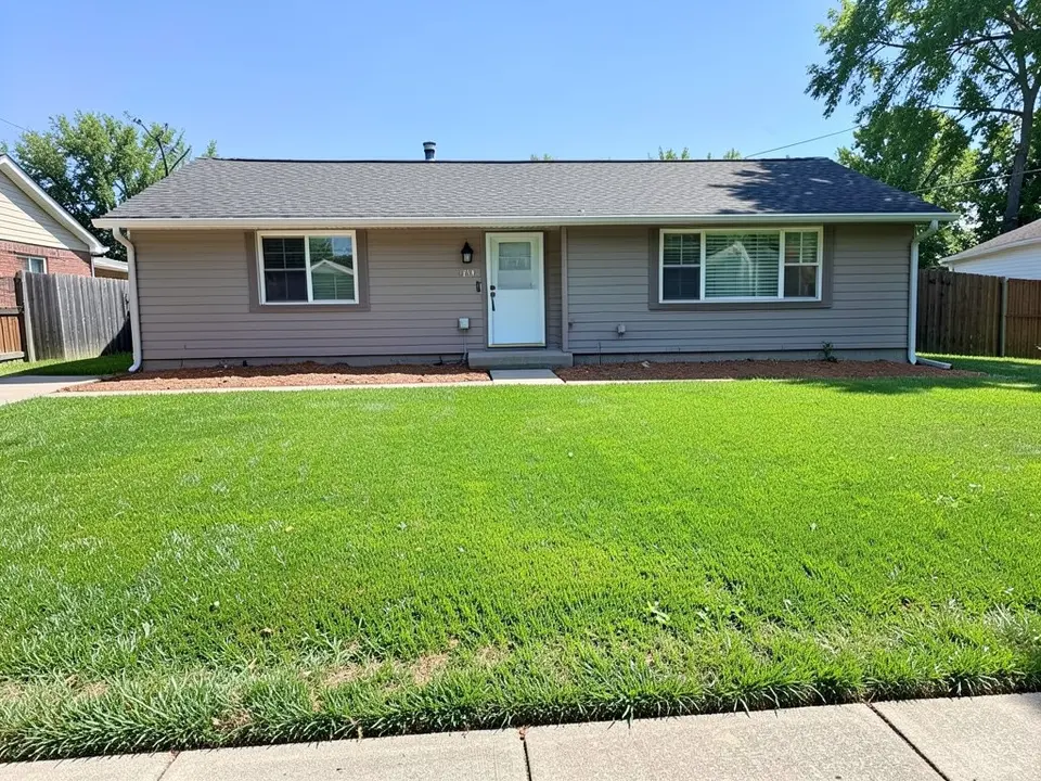 AI-generated example: the front of a grey one-story house with a white front door and two windows, a narrow strip of dark mulch along the house wall, a very large flat green lawn to the sidewalk and a wooden fence on each side