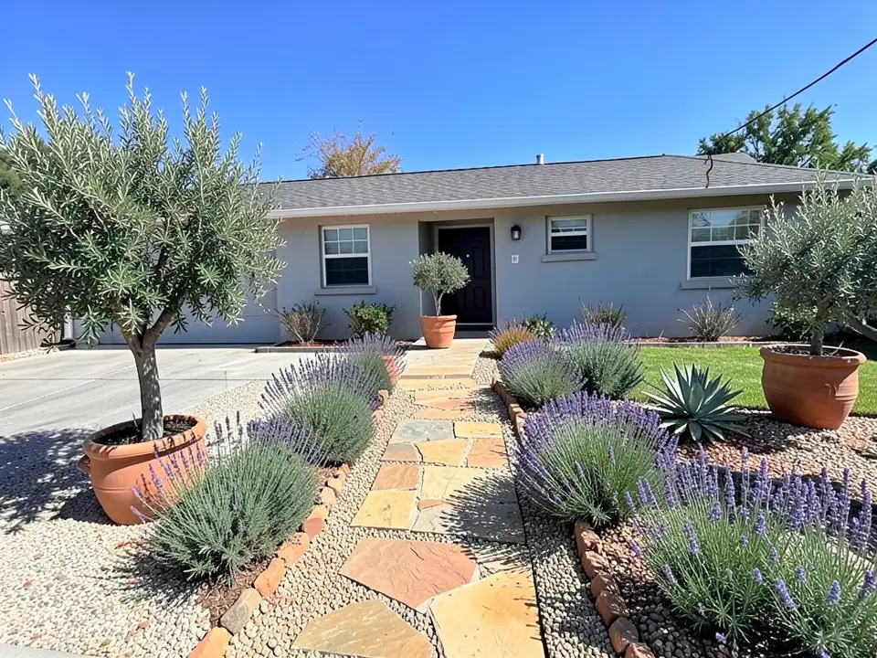 AI-edited version of the same image: the patchy lawn replaced by pebble gravel beds with lavender, an agave and terracotta-colored edging stones, a slightly curving flagstone path to the front door, olive trees in large terracotta pots on the left and right, a small potted olive by the door and a green lawn on the right; the front part of the driveway is now gravel