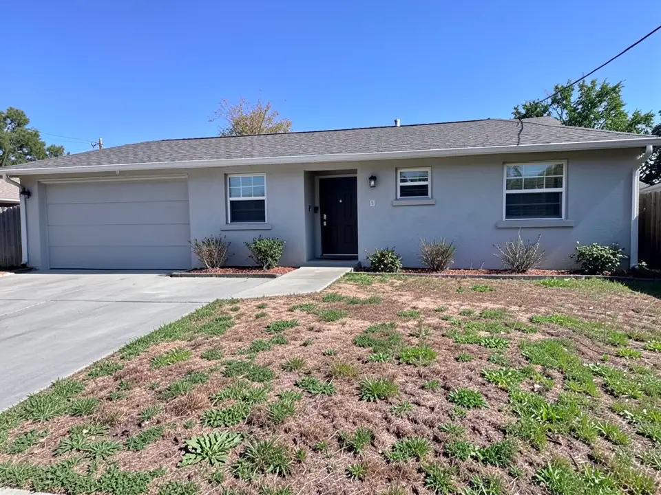 AI-generated example: a single-story light gray stucco ranch house with a garage, a dark front door and a few small shrubs along the wall, behind a patchy, half-bare front lawn, with a concrete driveway on the left and a concrete walk from the driveway to the front step