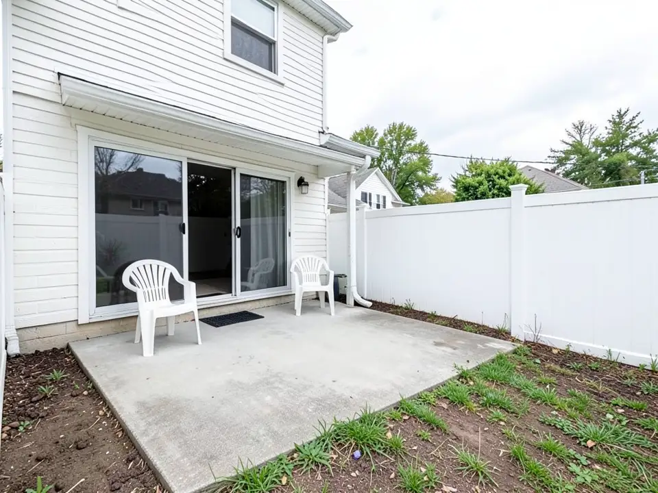 AI-generated example: a small gray concrete patio behind a white two-story house with a sliding glass door, two white plastic chairs and a doormat, next to a strip of bare soil and patchy grass along a white vinyl fence