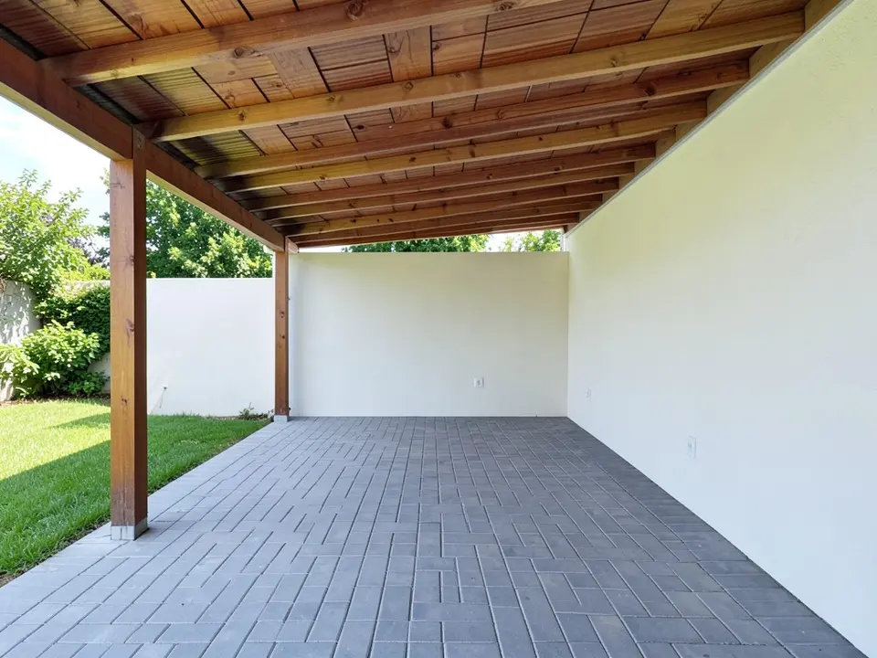 AI-generated example: a covered patio with a wooden slatted roof and grey paving, a plain white stucco wall at the far end, a lawn on the left and no furniture