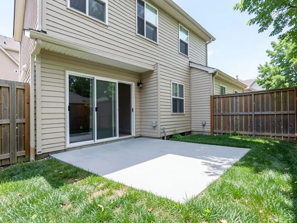 AI-generated example: the back of a beige two-story house with a sliding glass door, a plain grey concrete patio slab, a green lawn, a wooden fence on both sides and tree leaves at the top right
