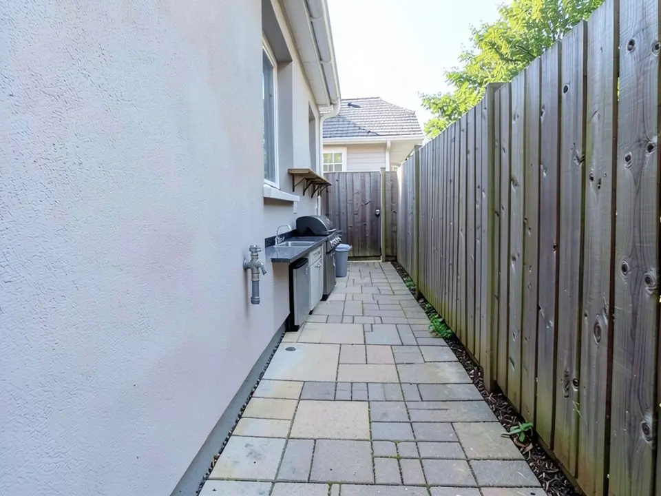 AI-edited version of the same image: a dark countertop run along the house wall in the distance with a sink, a black built-in grill, a stainless under-counter fridge, a small bracket shelf above it and a grey bin beside it, with the paved walkway still free next to it
