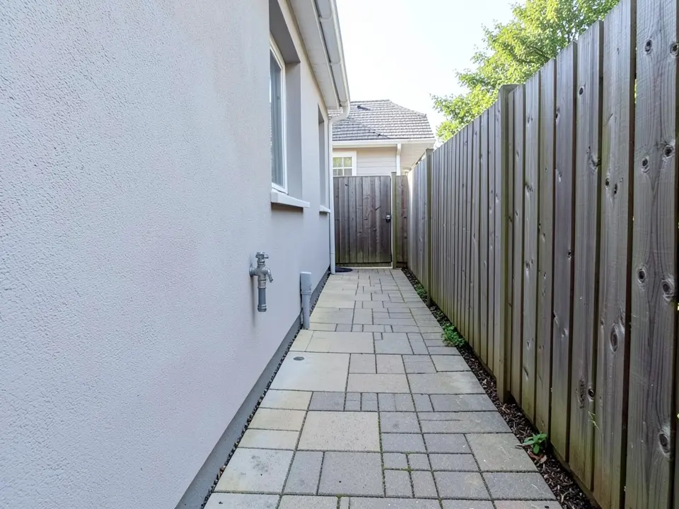 AI-generated example: a narrow side yard with a pale grey house wall on the left with a window and an outdoor tap, a paved path of grey and sand-colored pavers, a tall wooden fence on the right and a gate at the far end