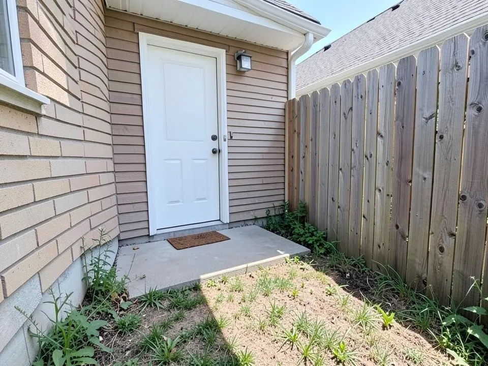AI-generated example: a narrow side corner of a house with a white door and a doormat on a small grey concrete slab, tan brick wall on the left, tan siding above, a tall wooden fence on the right, and bare dirt with weeds in front