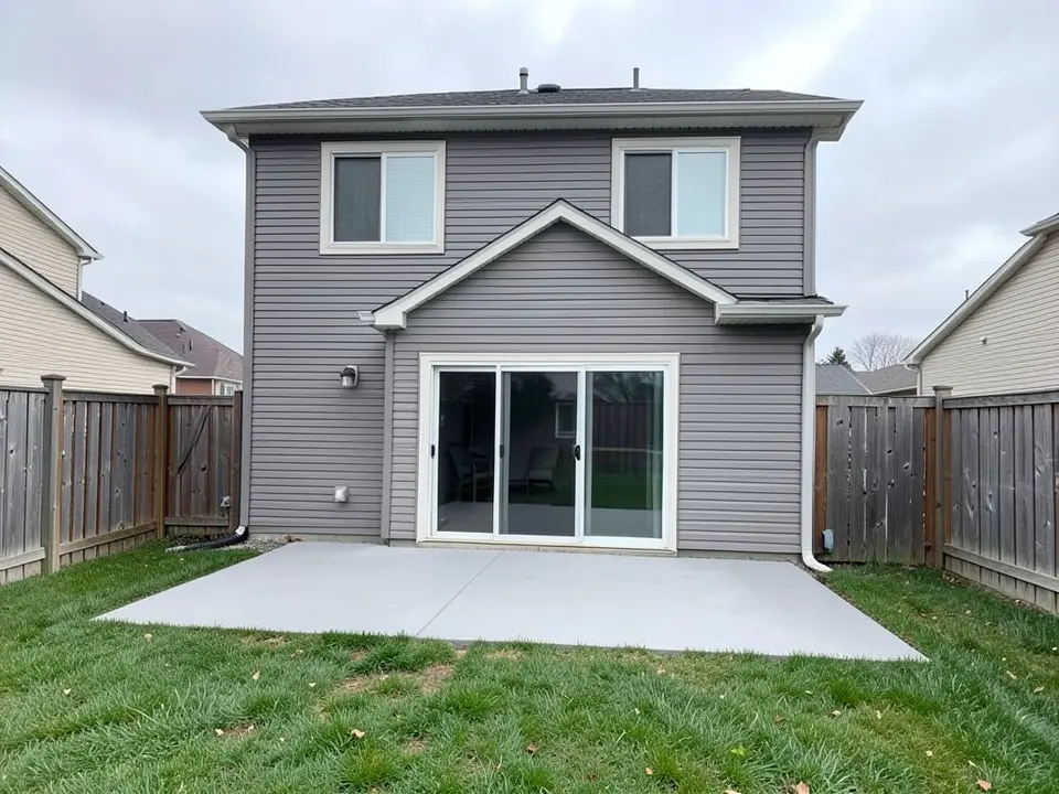 AI-generated example: the back of a grey two-story house with a gabled bump-out, a sliding glass door, a plain grey concrete patio slab, a green lawn and wooden fences on both sides