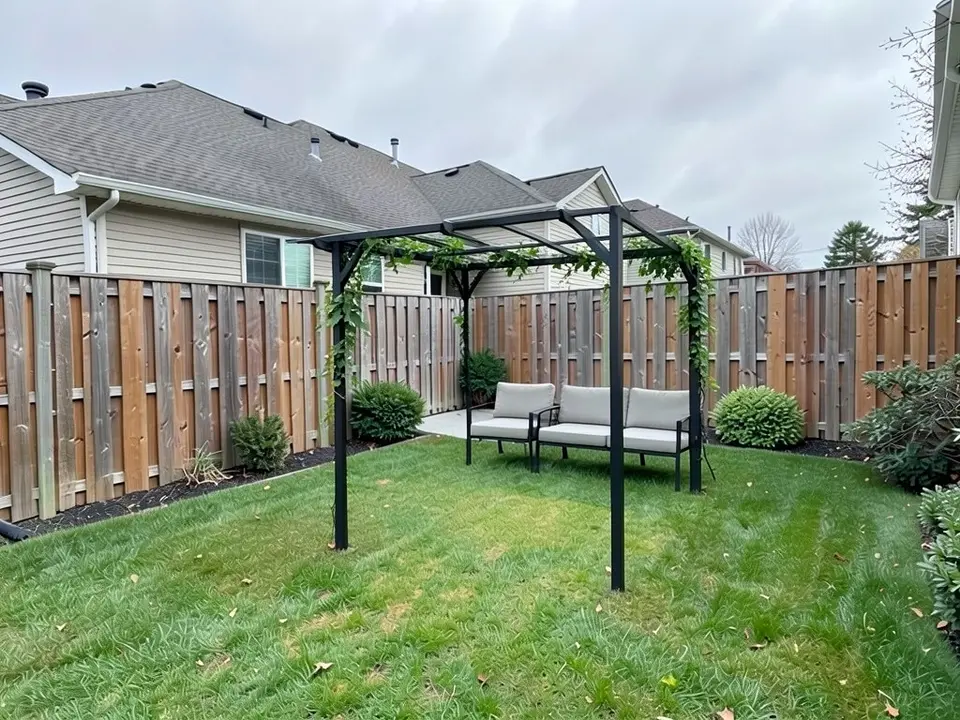 AI-edited version of the same image: a black metal pergola with open crossbeams stands on the lawn with a vine growing on it, and an outdoor sofa and a chair sit behind it by the fence