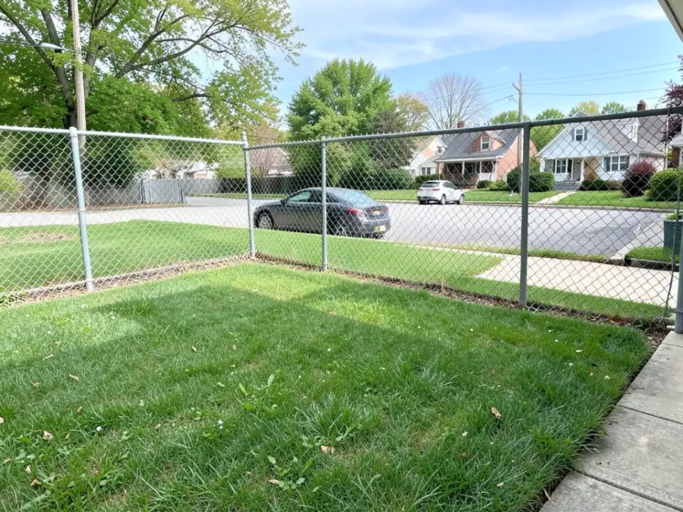 AI-generated example: a backyard lawn on a corner lot behind a gray chain-link fence, with a residential street, a parked dark sedan, a sidewalk and the houses across the street all in plain view