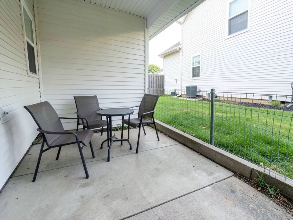 AI-generated example: a small covered concrete patio with three dark mesh chairs and a round metal table, open on the right to a neighbor's side yard behind a low green wire fence on a concrete curb, with the neighbor's white-sided house, an air-conditioning unit and lawn in full view