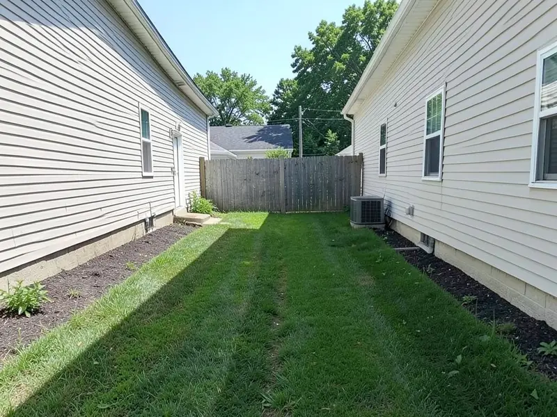 AI-generated example: a narrow strip of lawn between the side walls of two houses, with thin mulch strips along both walls, an air conditioner on the right and a wooden fence at the far end