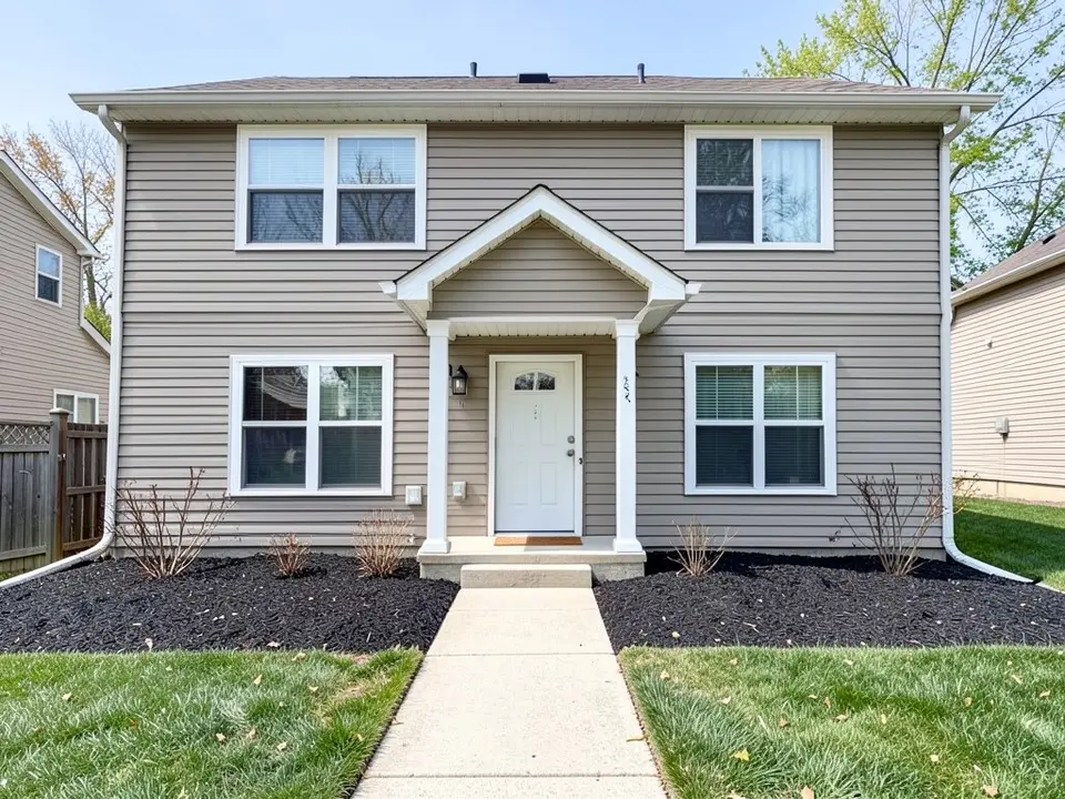 AI-generated example: the front of a two-story beige-siding house with a covered entry and white door, two windows either side and a long bed of dark mulch along the foundation with a few bare twiggy shrubs, a concrete walkway and lawn