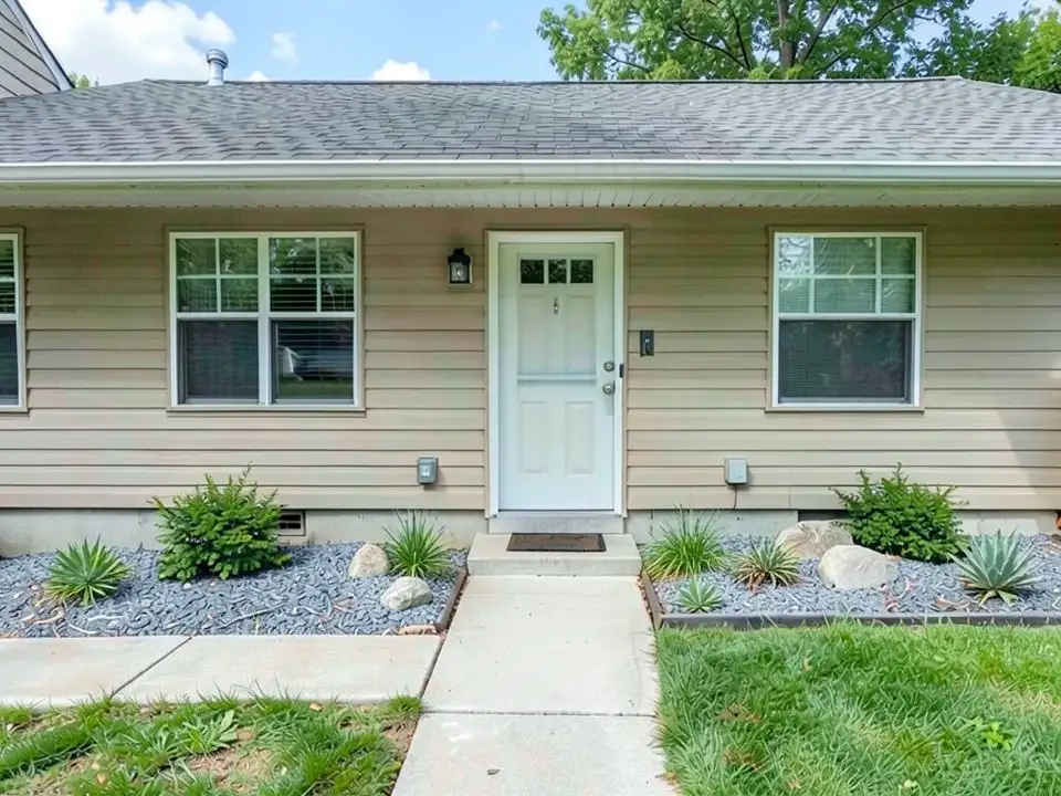 AI-edited version of the same image: the same house, door, path and lawn with the dark mulch replaced by grey gravel, a few small boulders and spiky grey-green plants, and the two green shrubs still in place