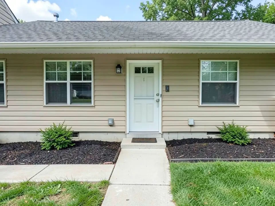 AI-generated example: the front of a one-story house with beige siding and a white front door, a concrete path to the door, a green lawn and two beds of dark wood-chip mulch along the foundation, each with one green shrub
