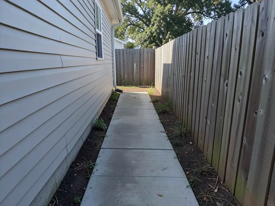 AI-generated example: a narrow side yard with light gray siding and a window on the left, a tall weathered wooden fence on the right, a straight concrete walkway between them and strips of bare dirt with a few weeds on both sides, a gate and a large tree at the far end